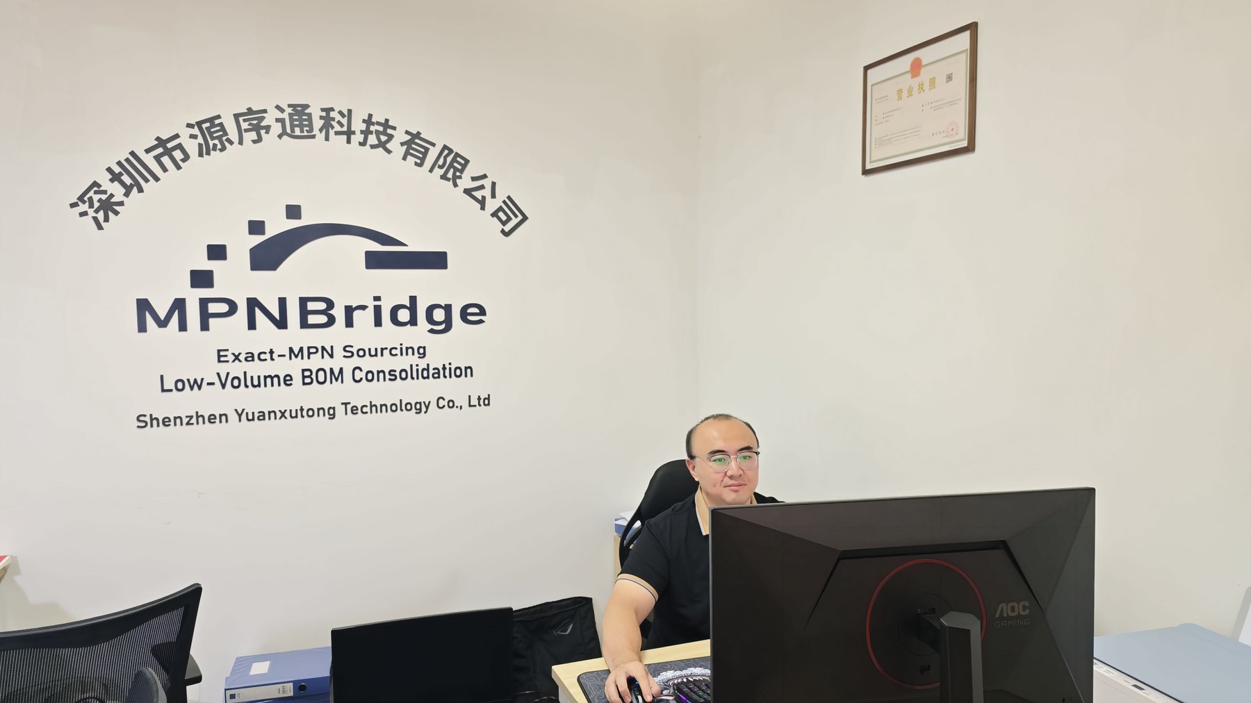 Jason Cheng working at the MPNBridge office in Shenzhen, in front of the company sign wall.